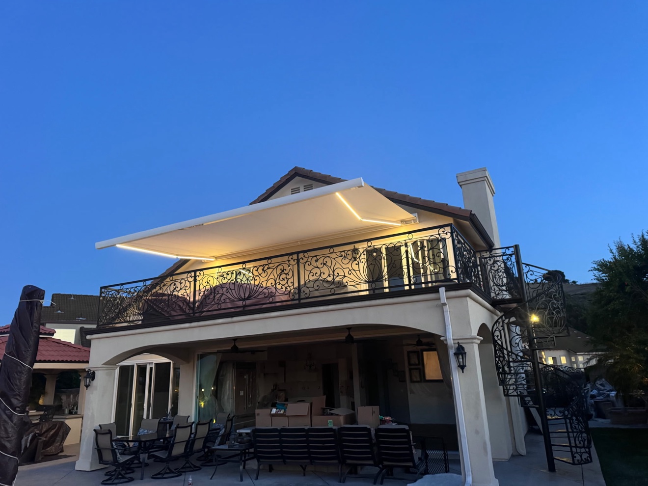 Wide view of a two-story home with LED-lit retractable awning over the balcony and spiral staircase