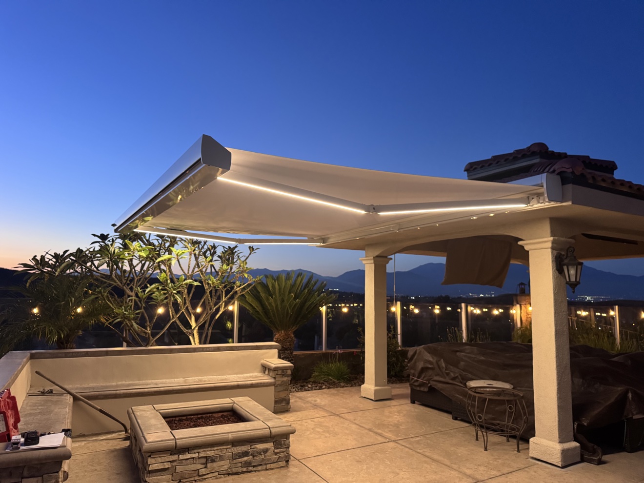 LED-lit retractable awning at dusk over a fire-pit patio with string lights and mountain views