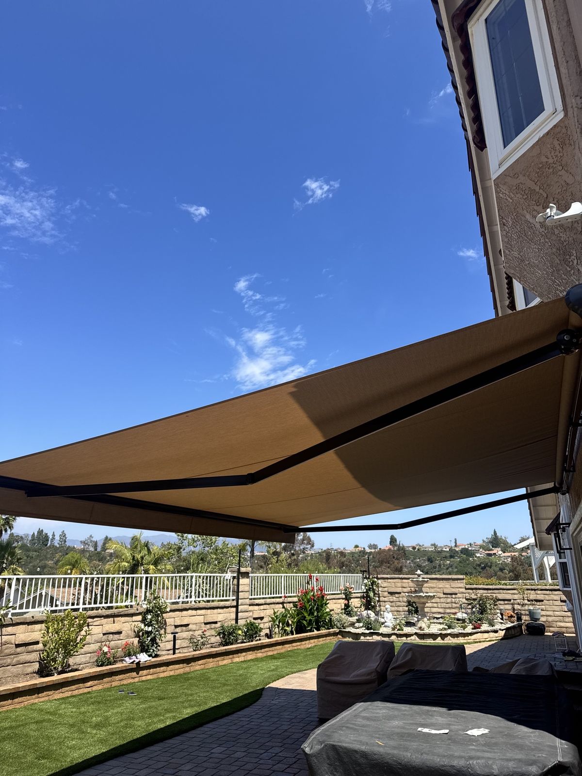 Tan retractable patio awning extending over a landscaped backyard with fountain and garden beds