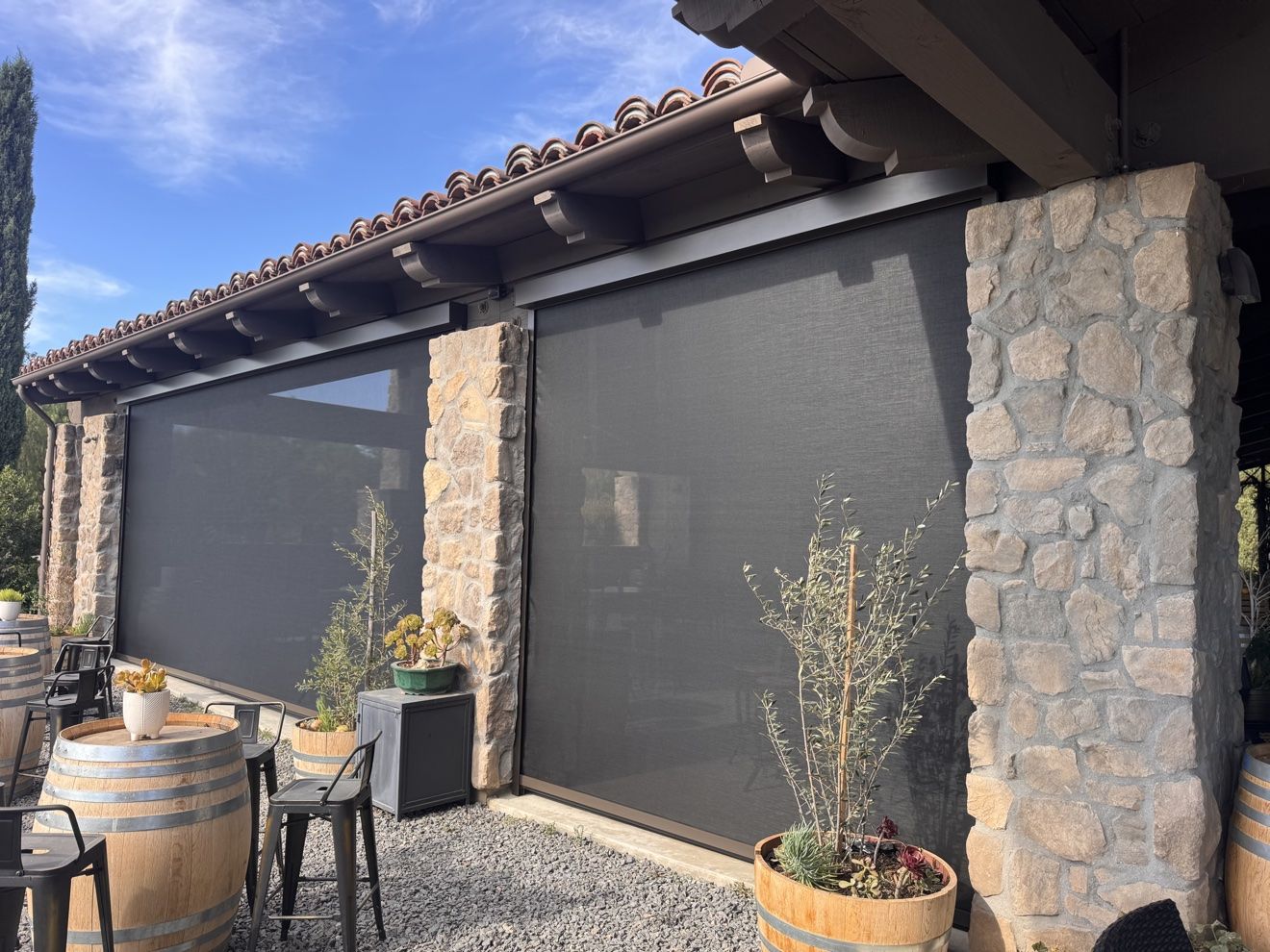Two large deployed motorized screens over a winery patio lined with barrels