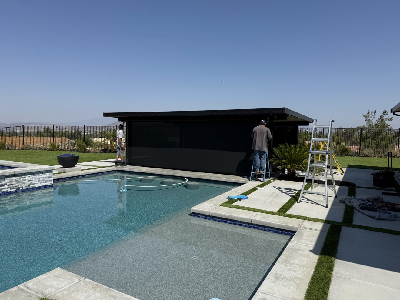 Two AJ Shades installers mounting a large motorized power screen beside a swimming pool