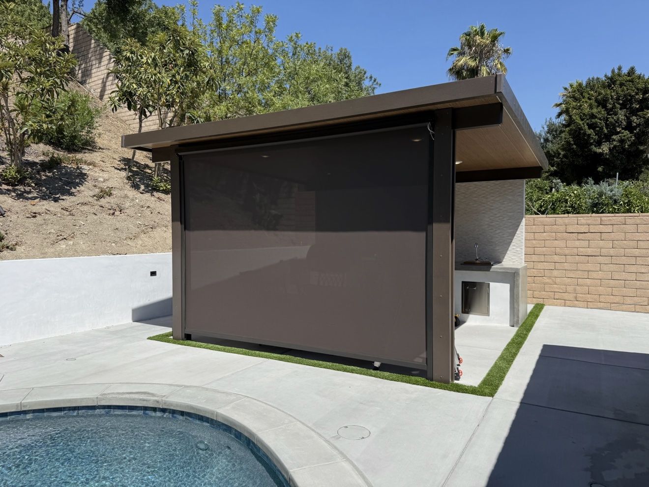 Dark motorized drop screen lowered on a poolside cabana structure with outdoor kitchen